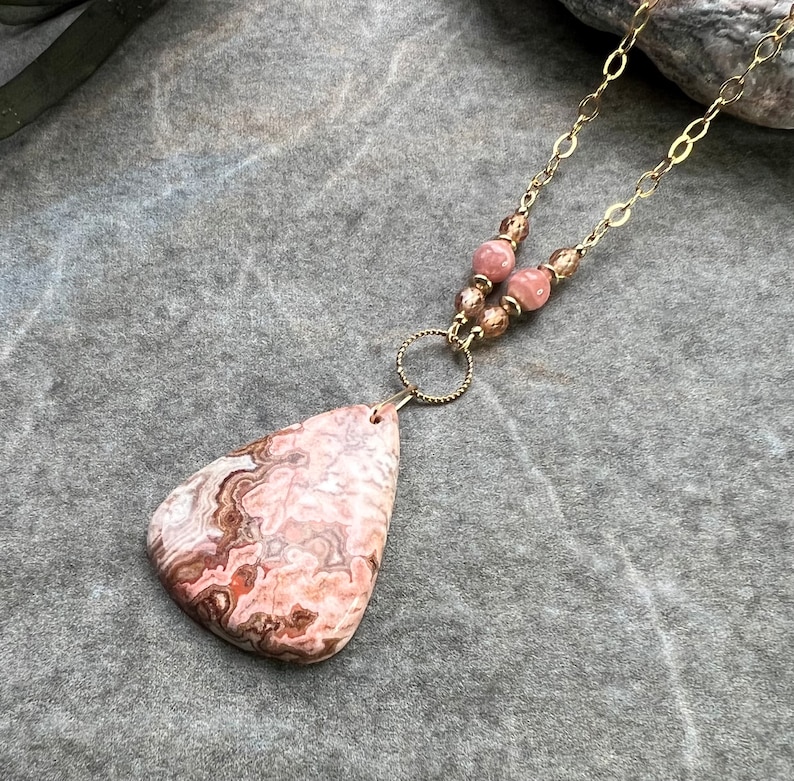 May include: A pink and brown agate pendant necklace with a gold chain and brown beads. The pendant is a teardrop shape with a swirling pattern.