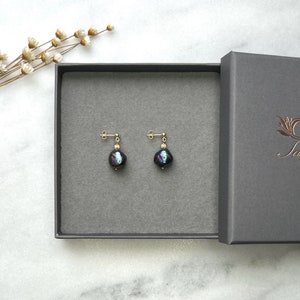 以下が含まれることがあります： イリデッセントブラックのバロックパールが付いた、ゴールドスタッドピアス1組。ダークグレーのギフトボックスに入っています。イヤリングは、グレーのベルベットのような表面に展示されています。箱は開いています。
