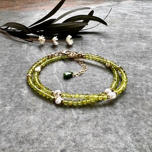 May include: A double-strand bracelet featuring faceted lime green peridot beads interspersed with small white freshwater pearls and gold-toned metal accents. The piece includes a gold-toned lobster clasp and a dangling dark green teardrop charm.