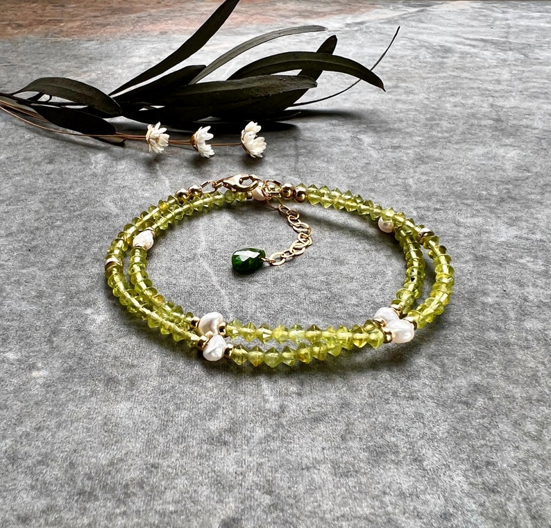 May include: A double-strand bracelet featuring faceted lime green peridot beads interspersed with small white freshwater pearls and gold-toned metal accents. The piece includes a gold-toned lobster clasp and a dangling dark green teardrop charm.