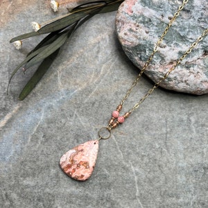 May include: A gold chain necklace with a pink and brown agate pendant and pink beads. The necklace is displayed on a grey textured surface with a rock in the background.