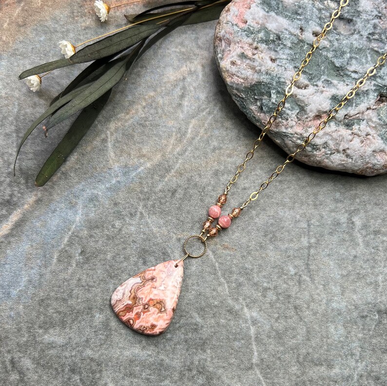 May include: A gold chain necklace with a pink and brown agate pendant and pink beads. The necklace is displayed on a grey textured surface with a rock in the background.