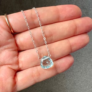 May include: A delicate silver chain necklace with a light blue gemstone pendant. The pendant is a faceted, teardrop-shaped stone.