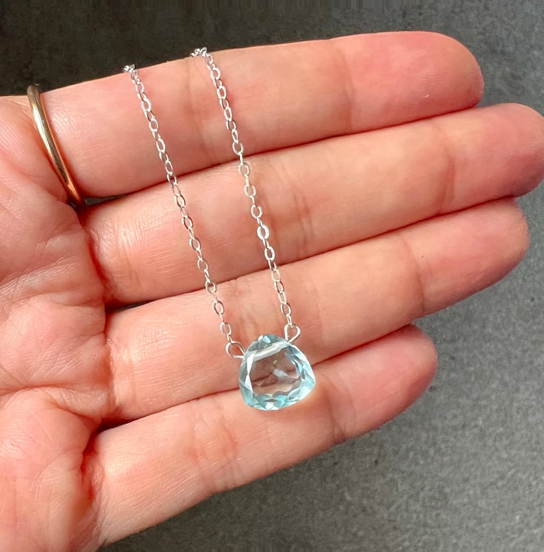 May include: A delicate silver chain necklace with a light blue gemstone pendant. The pendant is a faceted, teardrop-shaped stone.