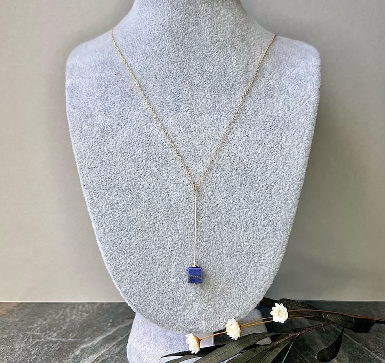 Puede incluir: Un collar de cadena dorada con un largo y delgado colgante que presenta una piedra cuadrada de lapisl&aacute;zuli azul oscuro.