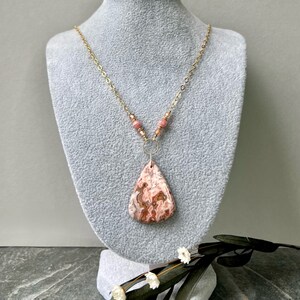 May include: A gold chain necklace with a pink and brown agate pendant and pink beads. The necklace is displayed on a grey mannequin.