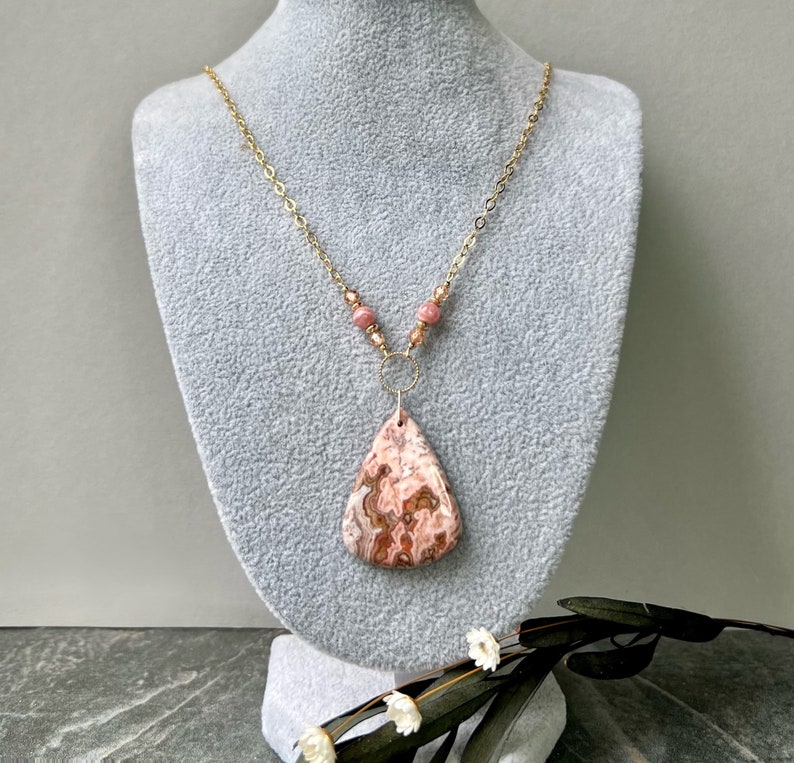 May include: A gold chain necklace with a pink and brown agate pendant and pink beads. The necklace is displayed on a grey mannequin.