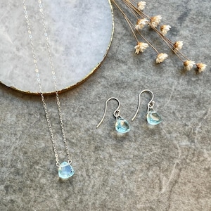 May include: A delicate silver necklace and matching earrings feature light blue gemstone pendants. The necklace has a simple chain and the earrings have a hook fastening.