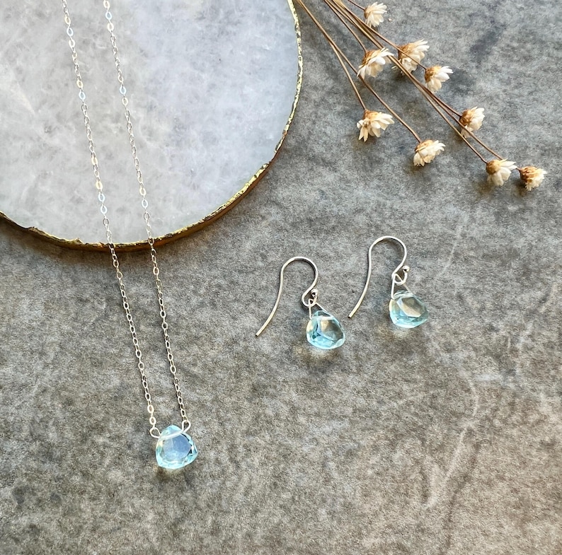 May include: A delicate silver necklace and matching earrings feature light blue gemstone pendants. The necklace has a simple chain and the earrings have a hook fastening.