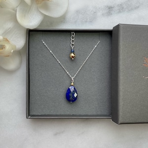 Könnte beinhalten: Eine silberne Halskette mit einem tropfenförmigen Anhänger aus blauem Lapislazuli und einem kleinen goldenen Perlenanhänger. Die Halskette wird in einer grauen Schmuckschatulle präsentiert.
