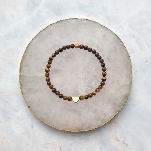 Könnte beinhalten: Ein braunes und goldenes Perlenarmband mit einem goldenen Herz-Anhänger. Das Armband liegt auf einem weißen Hintergrund.