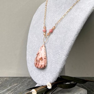 May include: A gold chain necklace with a pink and white teardrop-shaped pendant and pink beads. The necklace is displayed on a grey velvet mannequin.
