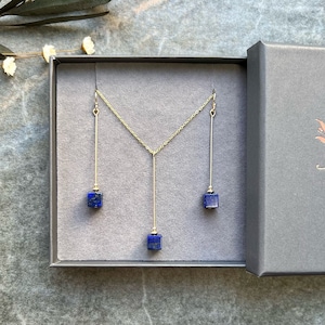 Puede incluir: Un collar y pendientes de oro con tres colgantes de piedra lapisl&aacute;zuli azul. Las joyas se muestran en una caja gris con una etiqueta blanca.