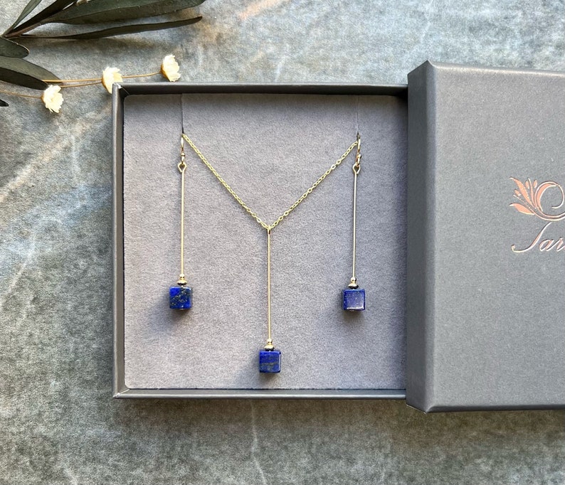 Puede incluir: Un collar y pendientes de oro con tres colgantes de piedra lapisl&aacute;zuli azul. Las joyas se muestran en una caja gris con una etiqueta blanca.
