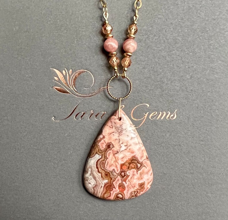 May include: A gold chain necklace with a pink and brown agate pendant and pink beads. The necklace is on a grey background with the text "Jara Gems".