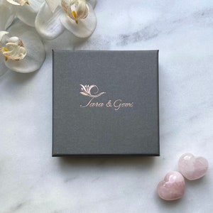 May include: A grey box with the text "Tara & Gems" in rose gold lettering. Two pink heart-shaped stones are on a white marble surface.