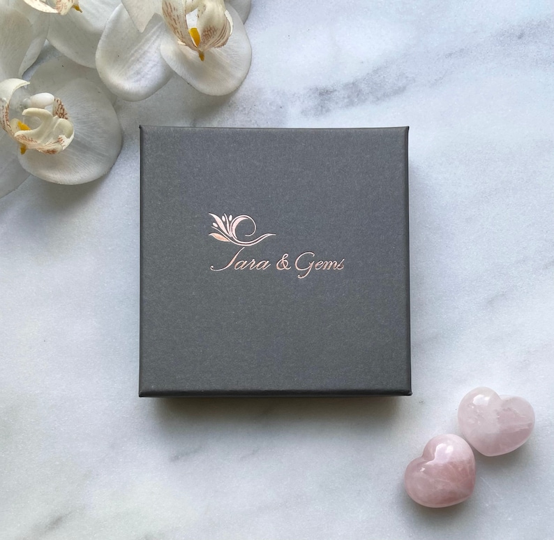 May include: A grey box with the text "Tara & Gems" in rose gold lettering. Two pink heart-shaped stones are on a white marble surface.