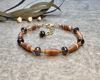 Pode incluir: Uma pulseira de contas com contas castanhas e bege em forma de barril, intercaladas com contas facetadas castanho escuro. A pulseira tem uma corrente e fecho dourados. As contas são enfiadas num cordão flexível.
