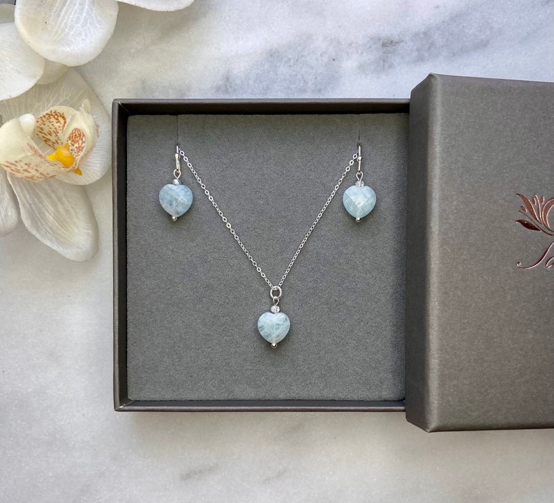 May include: A silver necklace and earrings set with light blue heart-shaped aquamarine stones. The jewellery is displayed in a grey box.