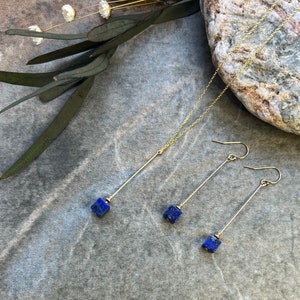 Puede incluir: Un collar y pendientes de oro con colgantes de piedra lapisl&aacute;zuli azul. El collar tiene una cadena larga y fina con un colgante cuadrado al final. Los pendientes son largos y finos con colgantes cuadrados en la parte inferior.