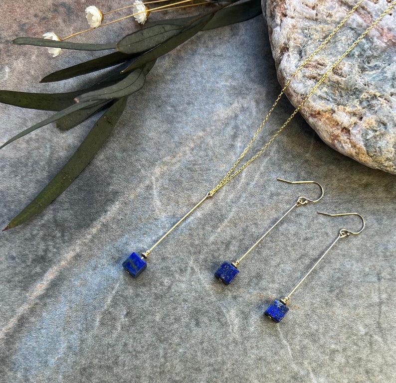 Puede incluir: Un collar y pendientes de oro con colgantes de piedra lapisl&aacute;zuli azul. El collar tiene una cadena larga y fina con un colgante cuadrado al final. Los pendientes son largos y finos con colgantes cuadrados en la parte inferior.