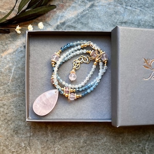 K&ouml;nnte beinhalten: Eine zarte Halskette mit einem tropfenf&ouml;rmigen Rosenquarz-Anh&auml;nger. Die Halskette hat hellblaue und klare Perlen, goldfarbenes Metall und rosa Edelsteine. Pr&auml;sentiert in einer grauen Geschenkbox.