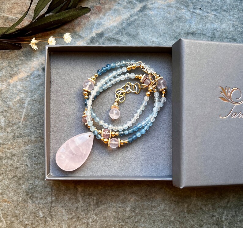 K&ouml;nnte beinhalten: Eine zarte Halskette mit einem tropfenf&ouml;rmigen Rosenquarz-Anh&auml;nger. Die Halskette hat hellblaue und klare Perlen, goldfarbenes Metall und rosa Edelsteine. Pr&auml;sentiert in einer grauen Geschenkbox.