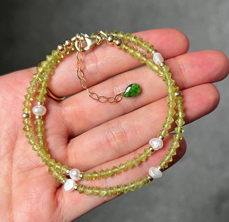 May include: A delicate bracelet featuring small, faceted lime-green peridot beads interspersed with white freshwater pearls and gold-toned metal spacers. The piece includes a gold-toned chain extender finished with a single, small, teardrop-shaped green gemstone charm.
