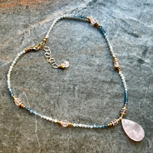 K&ouml;nnte beinhalten: Eine zarte Halskette mit kleinen, hellfarbenen Perlen und dunkleren blaugr&uuml;nen Perlen. Die Halskette enth&auml;lt goldfarbene Akzente und rosa Edelsteinperlen, mit einem gro&szlig;en, tropfenf&ouml;rmigen rosa Anh&auml;nger unten. Der Verschluss ist goldfarben.