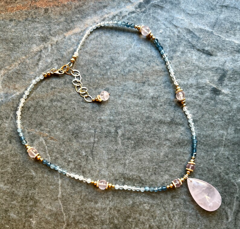 K&ouml;nnte beinhalten: Eine zarte Halskette mit kleinen, hellfarbenen Perlen und dunkleren blaugr&uuml;nen Perlen. Die Halskette enth&auml;lt goldfarbene Akzente und rosa Edelsteinperlen, mit einem gro&szlig;en, tropfenf&ouml;rmigen rosa Anh&auml;nger unten. Der Verschluss ist goldfarben.
