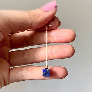 Puede incluir: Un pendiente largo y delgado de oro con una piedra cuadrada de lapisl&aacute;zuli azul oscuro en la parte inferior.