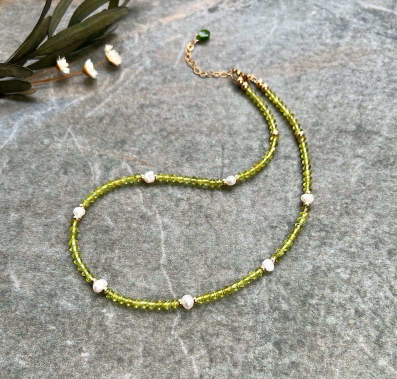 May include: A beaded necklace featuring small, faceted lime green peridot-colored crystals interspersed with irregular white freshwater pearls and tiny gold-toned metal spacers, finished with a gold-toned chain and a small green teardrop charm.