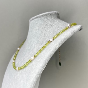 May include: A necklace featuring a strand of small, faceted lime green beads interspersed with irregular white pearls. The necklace includes a gold-toned metal chain extender at the end, finished with a small, dark green rectangular charm.