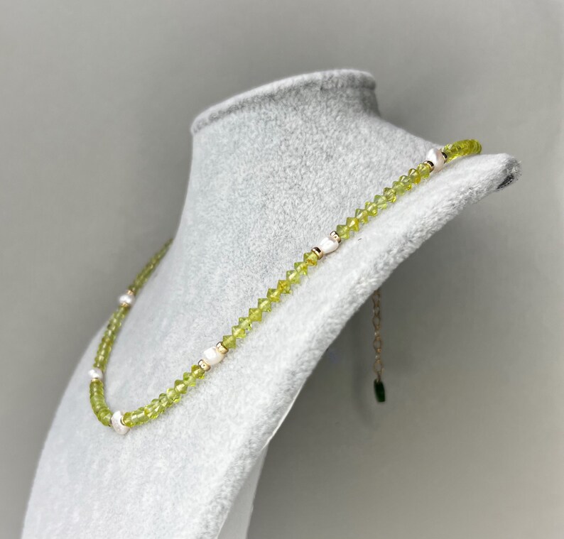 May include: A necklace featuring a strand of small, faceted lime green beads interspersed with irregular white pearls. The necklace includes a gold-toned metal chain extender at the end, finished with a small, dark green rectangular charm.