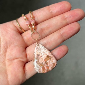 May include: A teardrop-shaped pendant necklace featuring a pink and brown agate stone. The necklace is crafted with gold-toned beads and a gold-toned chain.