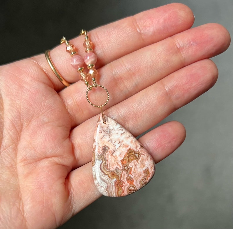 May include: A teardrop-shaped pendant necklace featuring a pink and brown agate stone. The necklace is crafted with gold-toned beads and a gold-toned chain.