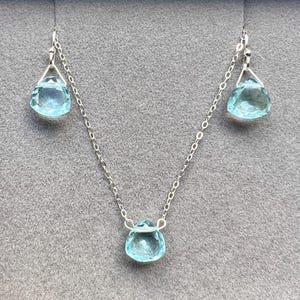 May include: A silver necklace with three teardrop-shaped blue gemstones. The necklace is on a grey background.