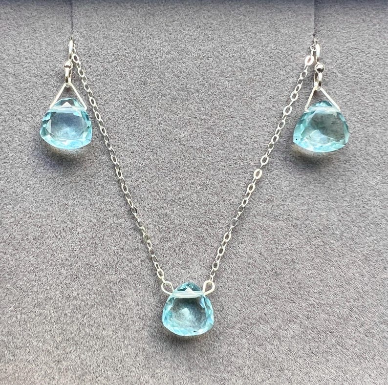 May include: A silver necklace with three teardrop-shaped blue gemstones. The necklace is on a grey background.