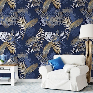 Puede incluir: Papel pintado azul marino con un diseño de hojas tropicales en dorado, blanco y azul. El patrón presenta diversas formas de hojas, creando un aspecto botánico denso. El papel pintado está en una pared detrás de un sillón blanco y una mesa auxiliar.