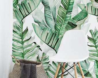 May include: A white chair with light wood legs sits in front of a wall covered in wallpaper featuring a pattern of large green banana leaves. A woven basket with a dark gray cloth draped over the side is positioned next to the chair.