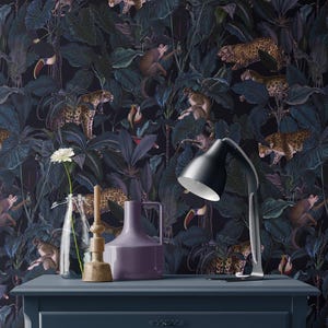 Op de afbeelding: Een donkerblauwe dressoir met een zwarte bureaulamp, een heldere glazen vaas met een witte bloem, een houten kandelaar en een paarse vaas. De achtergrond is een behang met een junglethema, met luipaarden, apen en toekans.