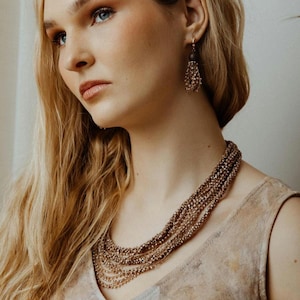 Può includere: Una collana a più fili con piccole perline color bronzo. La collana è indossata con orecchini abbinati. La modella indossa un top senza maniche di colore neutro. I gioielli sono di un tono caldo e terroso.