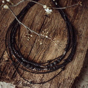 Può includere: Una collana multifilo realizzata con perline nere e cordoncino in pelle. La collana è a strati e presenta perline di diverse dimensioni, tra cui alcune con una superficie ruvida e testurizzata. La collana è esposta su una superficie in legno rustico.