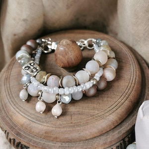 Op de afbeelding: Een armband met meerdere strengen, bestaande uit een mix van matte en gefacetteerde kralen in tinten crème, beige en zachtgrijs. Het ontwerp bevat kleine zilverkleurige metalen tussenstukjes, bungelende druppelvormige kralen en een zilverkleurige bedel met een gezicht.