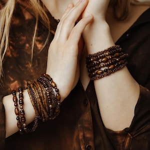 May include: Several wooden beaded bracelets in shades of brown. The bracelets have different bead shapes and sizes, creating a layered effect. The image shows the bracelets on a person's wrists.