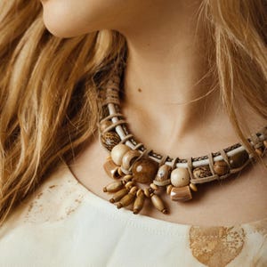 Pode incluir: Um colar artesanal com uma base de cordão trançado e contas de madeira em tons de castanho e bege. O colar tem uma estética tribal ou boêmia, com um aspeto natural e terroso.