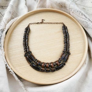 May include: A handcrafted wooden necklace with a layered design. The necklace features dark brown and tan wooden beads and rectangular shapes, strung together with a copper-coloured chain. The necklace is displayed on a light wooden tray.