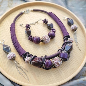 May include: A jewellery set comprising a necklace, bracelet, and earrings. The necklace and bracelet feature large, dark purple beads and smaller, textured beads. The earrings have a similar design with dangling silver chains. The set is displayed on a wooden tray.