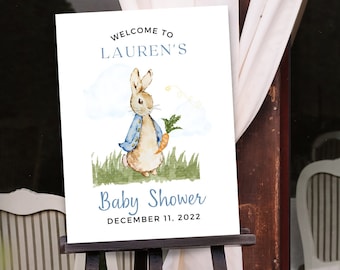 May include: Welcome to Lauren's Baby Shower sign with a watercolor illustration of Peter Rabbit holding a carrot. The sign is on a wooden easel and has a blue and white color scheme. The date of the shower is December 11, 2022.