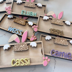 May include: A set of six wooden Easter bunny name tags with pink bunny ears, white clouds, and pink paw prints. Each tag has a different name and the text "Happy Easter".
